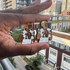 Op de afbeelding: Twee groene en gouden emaille pinnen van voetballers die shirts dragen met nummer 10. De spelers houden een voetbal vast en staan in een gooipositie.