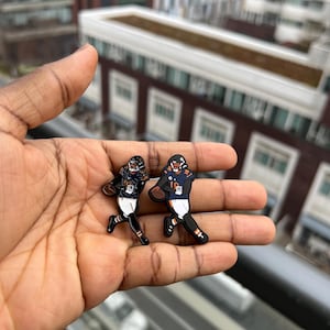 Könnte beinhalten: Zwei Emaille-Pins mit Chicago Bears Footballspielern in Action-Posen. Die Pins sind schwarz, orange und blau mit weißen Akzenten. Die Pins werden in einer Hand gehalten.