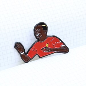 May include: A red and black enamel pin of a soccer player celebrating a goal. The player is wearing a red jersey with a yellow bird logo. The pin is on a white grid background.
