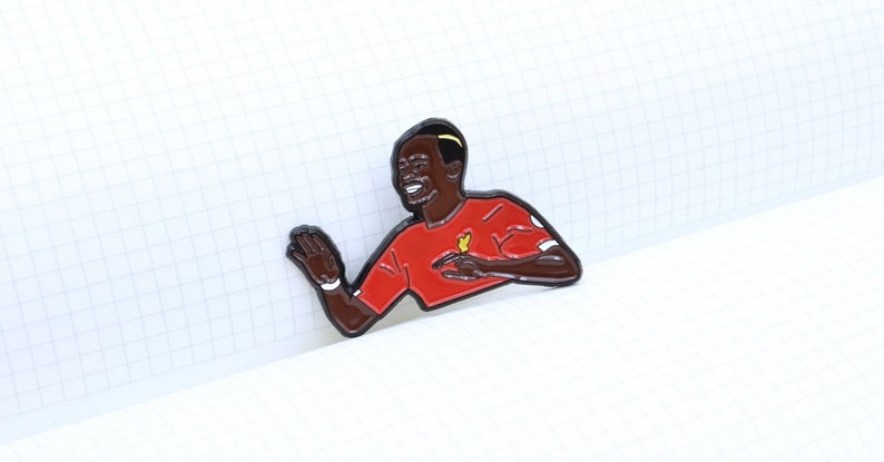 May include: A red and black enamel pin of a soccer player celebrating a goal. The player is wearing a red jersey with a yellow bird logo. The pin is on a white grid background.