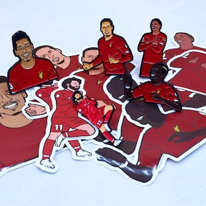 May include: A collection of red and black enamel pins featuring different soccer players. The pins are on a white background and are arranged in a random pattern.