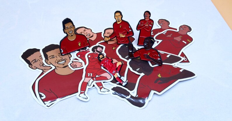 May include: A collection of red and black enamel pins featuring different soccer players. The pins are on a white background and are arranged in a random pattern.