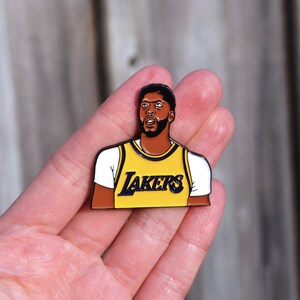 Puede incluir: Un pin de esmalte amarillo y morado con un retrato de dibujos animados de un jugador de baloncesto que lleva una camiseta de los Lakers. El jugador est&aacute; mirando hacia arriba y tiene una expresi&oacute;n seria en su rostro.