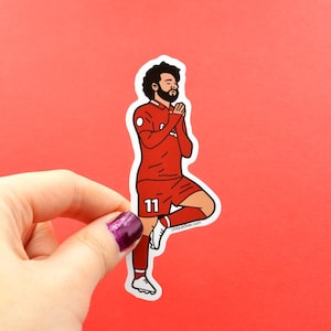 May include: A red and white soccer player sticker with the number 11 on the jersey. The player is in a praying pose with their arms crossed in front of their chest. The sticker is on a red background.