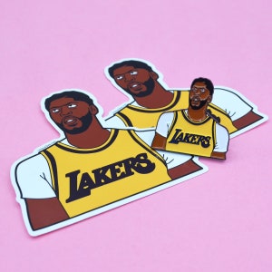 Puede incluir: Dos pegatinas de jugadores de baloncesto de los Lakers, amarillas y negras, con la palabra "LAKERS" impresa en la parte delantera. Tambi&eacute;n se muestra una pegatina m&aacute;s peque&ntilde;a y similar de un jugador de los Lakers.
