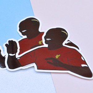 May include: Two red and white stickers of a cartoon person with a brown skin tone, black hair, and a yellow headband. The person is wearing a red shirt with a yellow symbol on the left side of the chest. The person is smiling and has their arms outstretched.