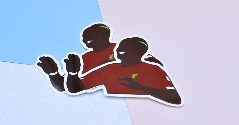 May include: Two red and white stickers of a cartoon person with a brown skin tone, black hair, and a yellow headband. The person is wearing a red shirt with a yellow symbol on the left side of the chest. The person is smiling and has their arms outstretched.