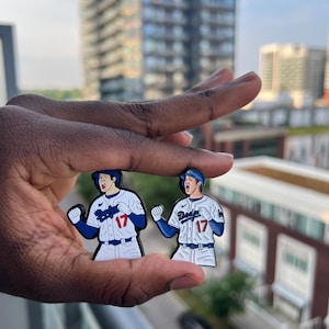 Könnte beinhalten: Zwei Baseballspieler-Anstecknadeln. Jede Anstecknadel zeigt einen Spieler in einer weiß-blauen Uniform mit der Nummer 17. Die Anstecknadeln haben ein cartoonartiges Design, und das Wort "Dodgers" ist auf der Uniform zu sehen.