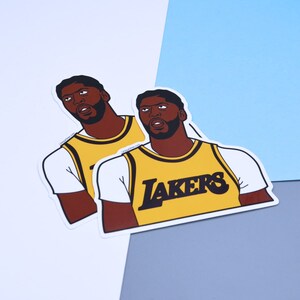 Puede incluir: Dos pegatinas de un jugador de baloncesto que lleva una camiseta amarilla con la palabra "LAKERS" impresa en la parte delantera. El jugador tiene un tono de piel negro y marr&oacute;n y est&aacute; mirando hacia arriba.