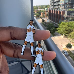 Puede incluir: Dos pines de esmalte con una ilustración de dibujos animados de un jugador de fútbol con una camiseta blanca con detalles en negro y dorado. El jugador lleva pantalones cortos blancos y calcetines negros. El jugador está haciendo un gesto con las dos manos. Los pines están en una mano.