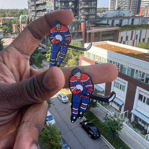 May include: Two enamel pins depicting a hockey player in a blue and red uniform with a white helmet. The player is holding a hockey stick and is in a skating position. The pins are held in a hand.