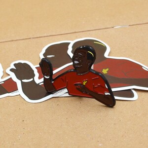 May include: A black and red enamel pin of a smiling soccer player wearing a red jersey with a yellow star. The player is raising his right hand in the air.