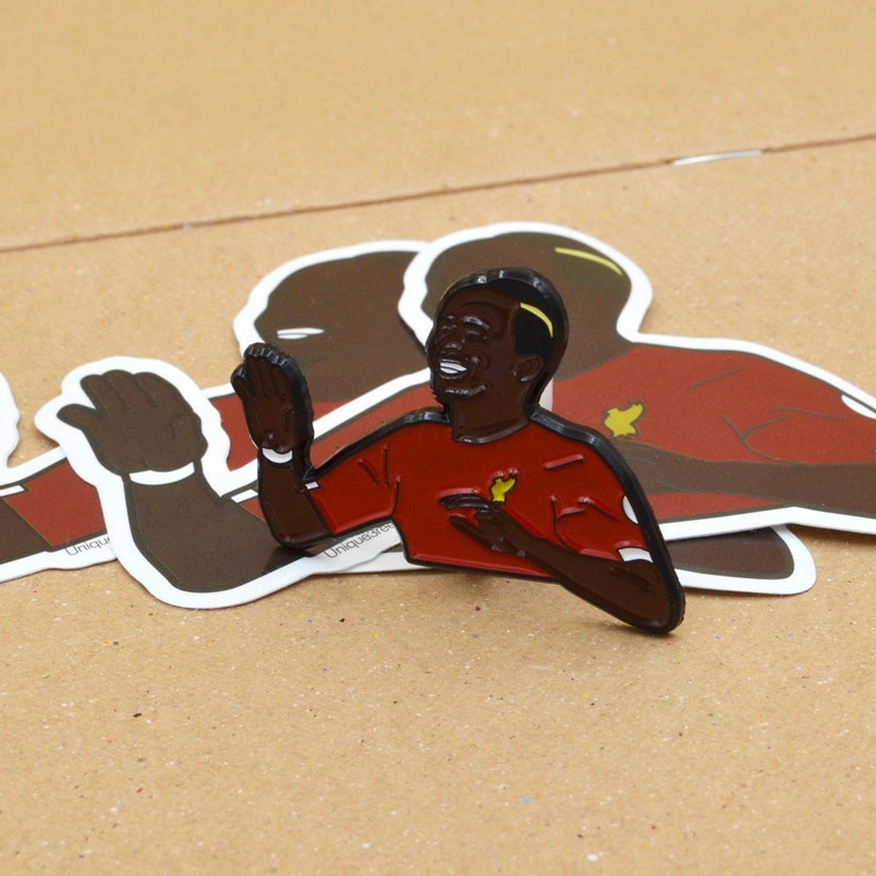 May include: A black and red enamel pin of a smiling soccer player wearing a red jersey with a yellow star. The player is raising his right hand in the air.