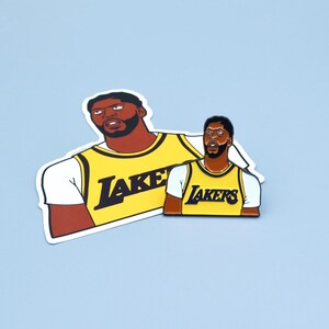 Puede incluir: Una camiseta de baloncesto amarilla y negra con la palabra "LAKERS" impresa en la parte delantera. La camiseta es un bot&oacute;n de solapa.