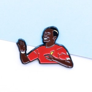 May include: A red and black enamel pin featuring a cartoon illustration of a soccer player celebrating a goal. The player is wearing a red jersey with a yellow bird logo.
