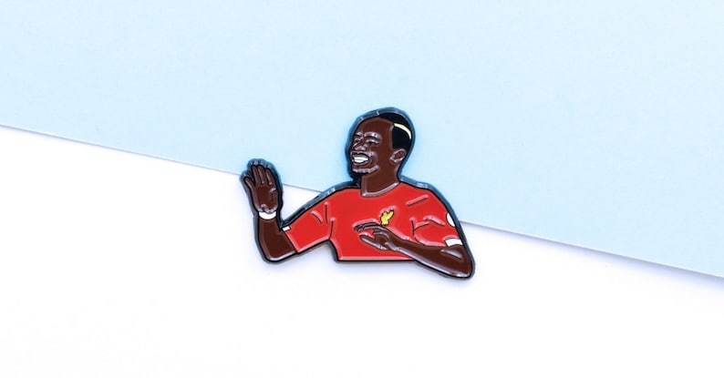 May include: A red and black enamel pin featuring a cartoon illustration of a soccer player celebrating a goal. The player is wearing a red jersey with a yellow bird logo.