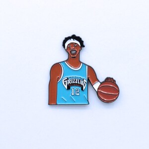 Puede incluir: Un pin de esmalte azul y blanco con un jugador de baloncesto que lleva una camiseta azul con el texto "Vancouver Grizzlies" y el número "12". El jugador está sosteniendo un balón de baloncesto.