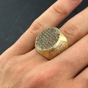May include: A gold-toned signet ring featuring an oval face with intricate, deeply engraved Arabic calligraphy. The band also displays detailed script along its sides, creating a textured, metallic appearance with a polished finish.