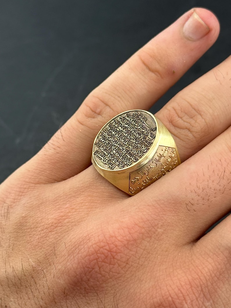 May include: A gold-toned signet ring featuring an oval face with intricate, deeply engraved Arabic calligraphy. The band also displays detailed script along its sides, creating a textured, metallic appearance with a polished finish.