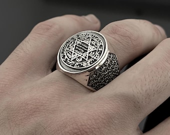 Op de afbeelding: Een zilveren ring met een gedetailleerd, cirkelvormig ontwerp met een zespuntige ster en ingewikkelde patronen. De ring heeft een brede band met een textuurpatroon. De ring wordt aan een vinger gedragen.