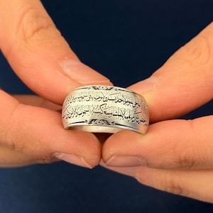 May include: A silver ring with engraved Arabic script. The ring is wide and has a polished finish. The text is in black and wraps around the entire band. The ring is held between two hands against a dark blue background.