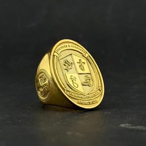 Könnte beinhalten: Ein goldfarbener Siegelring mit ovaler Front und einem eingravierten Wappen, das in vier Quadranten unterteilt ist. Die Schriftzüge "FOUNDER & OVERSEER", "FRESH SPRINGS MINISTRIES" und "ANDRE D. LEE" sind um das zentrale Schildmotiv eingraviert.
