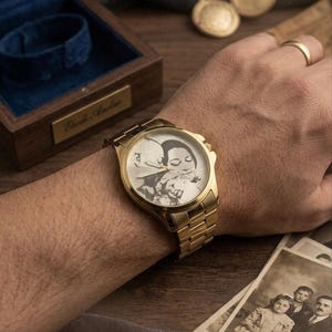 Puede incluir: Un reloj de pulsera dorado con una imagen grabada personalizada en la esfera. El reloj est&aacute; en una mu&ntilde;eca, con una correa dorada y una esfera rectangular. Una caja de madera y fotograf&iacute;as antiguas est&aacute;n en el fondo.