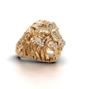 Op de afbeelding: Een gouden ring in de vorm van een leeuwenkop met gedetailleerde manen en gelaatstrekken. Het voorhoofd en de snuit van de leeuw zijn geaccentueerd met kleine, heldere, rondgeslepen steentjes die schitteren op het gepolijste gouden oppervlak.