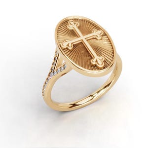 Könnte beinhalten: Ein goldener Ring mit ovaler Platte, die ein erhabenes Kreuzemblem zeigt, umgeben von gravierten Sonnenstrahlen. Die Ringschiene teilt sich oben in zwei Arme, die jeweils mit einer Reihe kleiner, klarer, runder Steine besetzt sind.