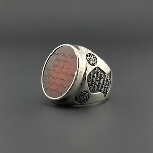 Puede incluir: Un anillo de plata con una gran piedra ovalada de color marrón rojizo grabada con caligrafía árabe. La banda de metal está decorada con intrincadas inscripciones árabes adicionales y motivos circulares en los laterales, acabado pulido.