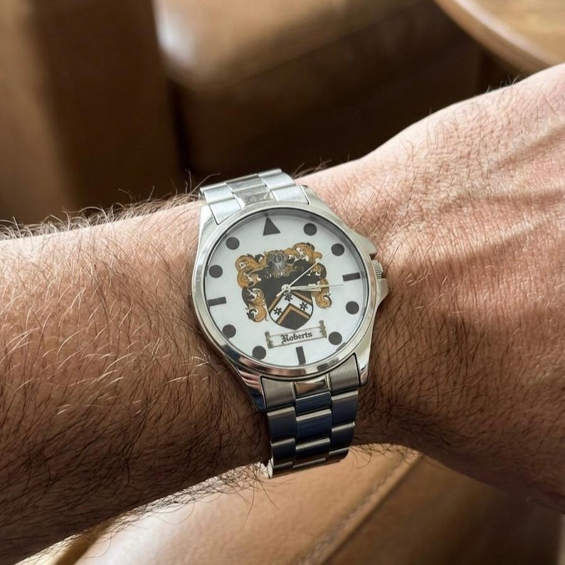 May include: A silver wristwatch with a white face featuring a family crest design and the name "Roberts" below. The watch has a silver band and is worn on a wrist.