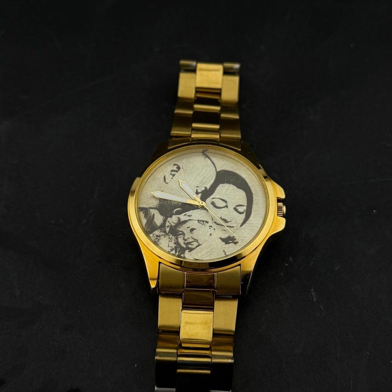 Puede incluir: Reloj de pulsera dorado con una imagen detallada en blanco y negro en la esfera. El reloj tiene una correa dorada y una esfera redonda con un retrato de una persona. Un accesorio cl&aacute;sico.