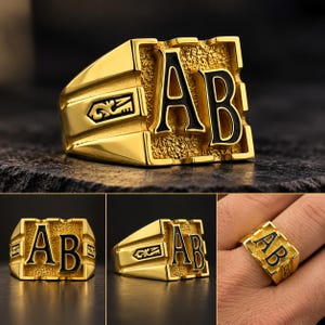 May include: A gold-toned signet ring featuring the black-filled letters AB on a textured rectangular face. The sides of the ring display a decorative engraved symbol. The design is bold with a polished finish on the band and edges.