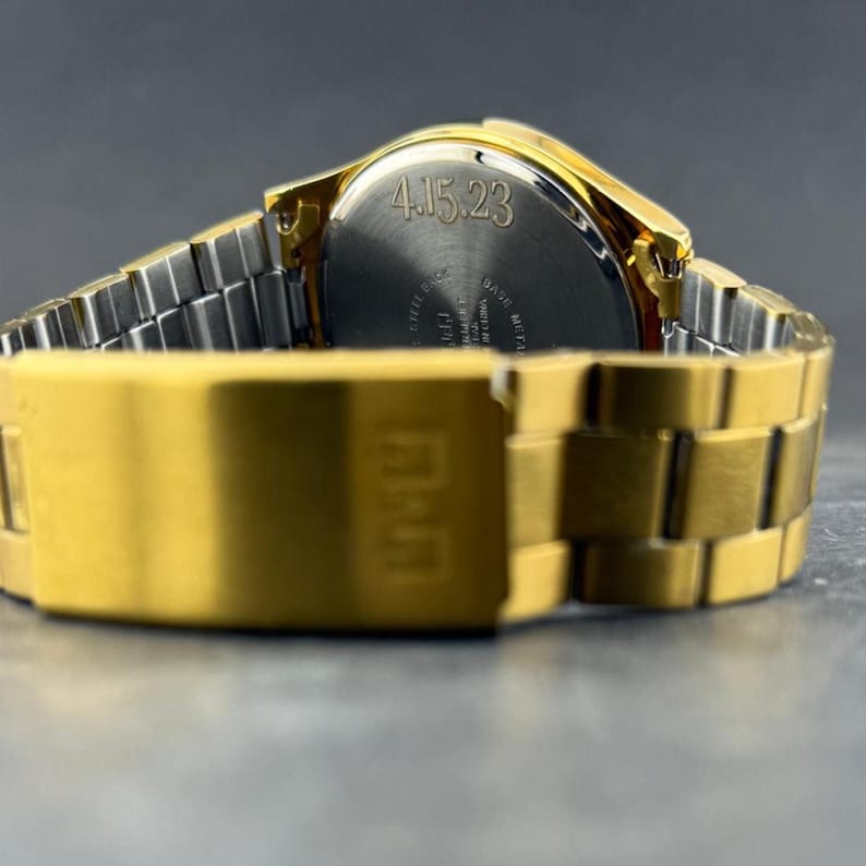 May include: Close-up of a gold-toned wristwatch with a silver band. The watch face displays the date "4.15.23". The watch band and clasp are in focus, showcasing the gold color. The watch is on a dark surface.
