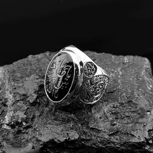 May include: A silver ring with an oval black stone, featuring Arabic calligraphy. The ring has intricate side detailing and a polished finish. The ring is displayed on a textured, dark gray surface.