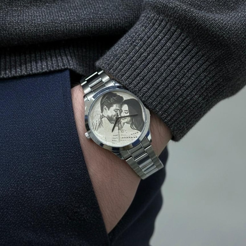 May include: Silver wristwatch with a stainless steel band. The watch face features a black and white image of a couple. The watch is on a wrist, with a gray sweater and navy blue pants in the background.