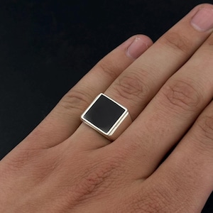 May include: A silver ring with a square, black onyx stone. The ring has a polished finish and is shown on a person's finger. The black stone is set within a silver bezel.