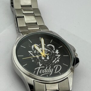 May include: Silver-toned wristwatch with a black watch face. The face features a bear graphic wearing glasses, with the text "Teddy D" below. The watch has a metal band and gold-colored hands.