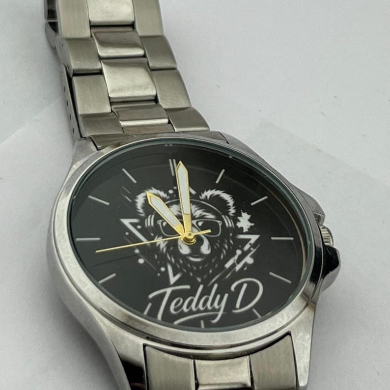 May include: Silver-toned wristwatch with a black watch face. The face features a bear graphic wearing glasses, with the text "Teddy D" below. The watch has a metal band and gold-colored hands.
