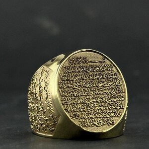 May include: A gold-toned ring featuring an oval face engraved with intricate Arabic calligraphy. The band also displays detailed script carvings along its sides, creating a textured appearance across the entire surface of the metal jewelry piece.