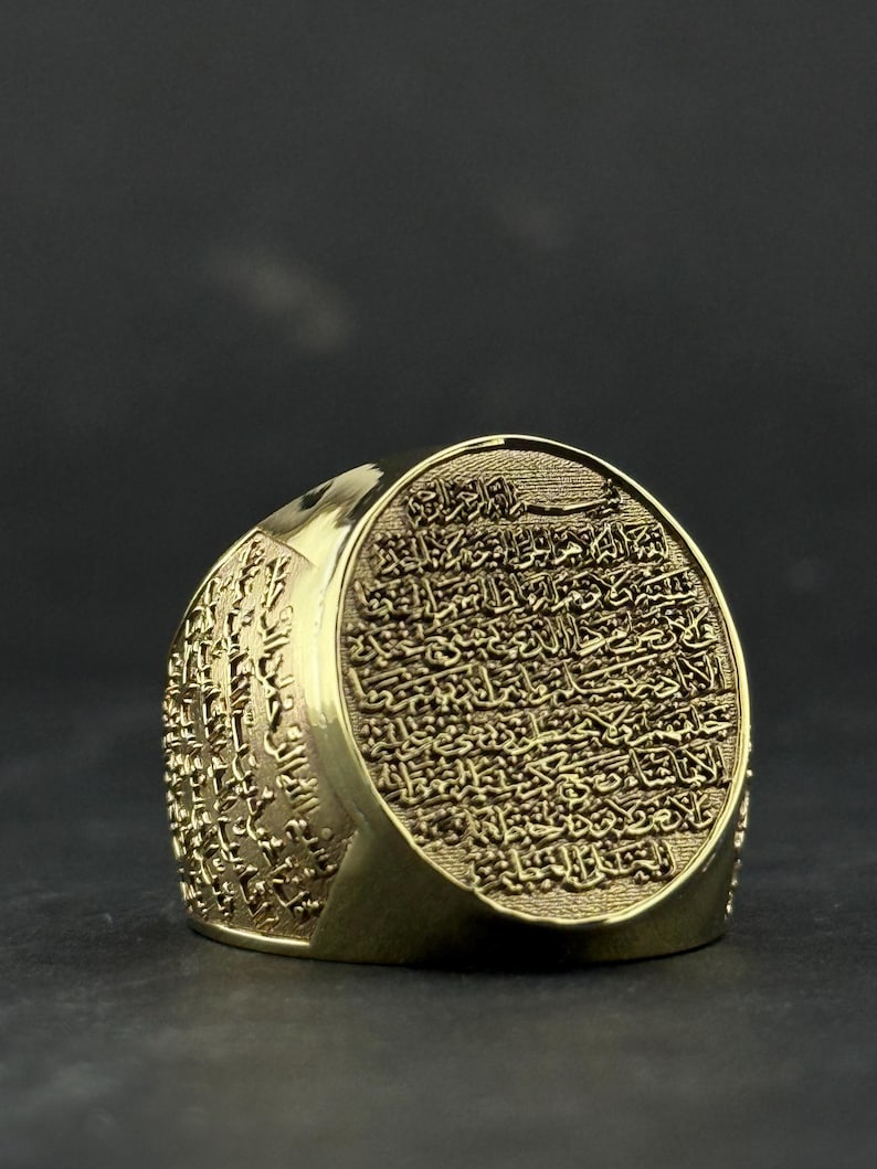 May include: A gold-toned ring featuring an oval face engraved with intricate Arabic calligraphy. The band also displays detailed script carvings along its sides, creating a textured appearance across the entire surface of the metal jewelry piece.