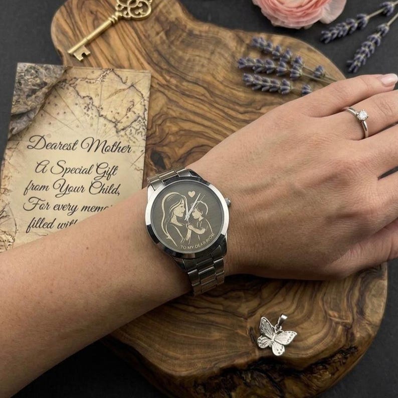 K&ouml;nnte beinhalten: Eine silberne Armbanduhr mit einem Armband aus Edelstahl. Das Zifferblatt zeigt ein eingraviertes Bild einer Mutter und eines Kindes. Die Uhr befindet sich an einem Handgelenk, mit einem Ring und einem Schmetterlingsanh&auml;nger. Eine Karte mit Text ist ebenfalls im Bild.