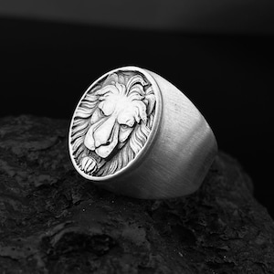 Puede incluir: Un anillo de plata con un diseño detallado de cabeza de león. El anillo está sobre una superficie oscura y texturizada.