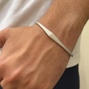 May include: A silver-colored bracelet with a geometric design. The bracelet has a flat, elongated center and tapers to a thin band on each side. The jewelry is worn on a wrist.