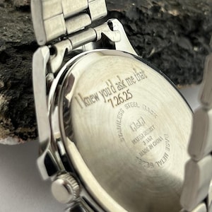 May include: Silver stainless steel watch with a rectangular face and a metal band. The watch back is engraved with the text "I knew you'd ask me that 7.26.25". The watch is water resistant and made in China.