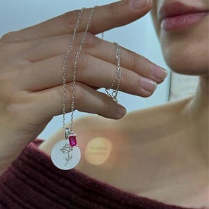 May include: A silver necklace with a round pendant featuring a delicate flower design and a small pink gemstone. The pendant also has the words "Endless Journey" engraved on it.