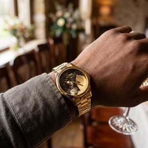 Puede incluir: Un reloj de pulsera dorado con una esfera detallada que presenta una imagen en blanco y negro. El reloj tiene una correa dorada y se lleva en la mu&ntilde;eca de una persona. El reloj es el punto focal, con un fondo borroso.