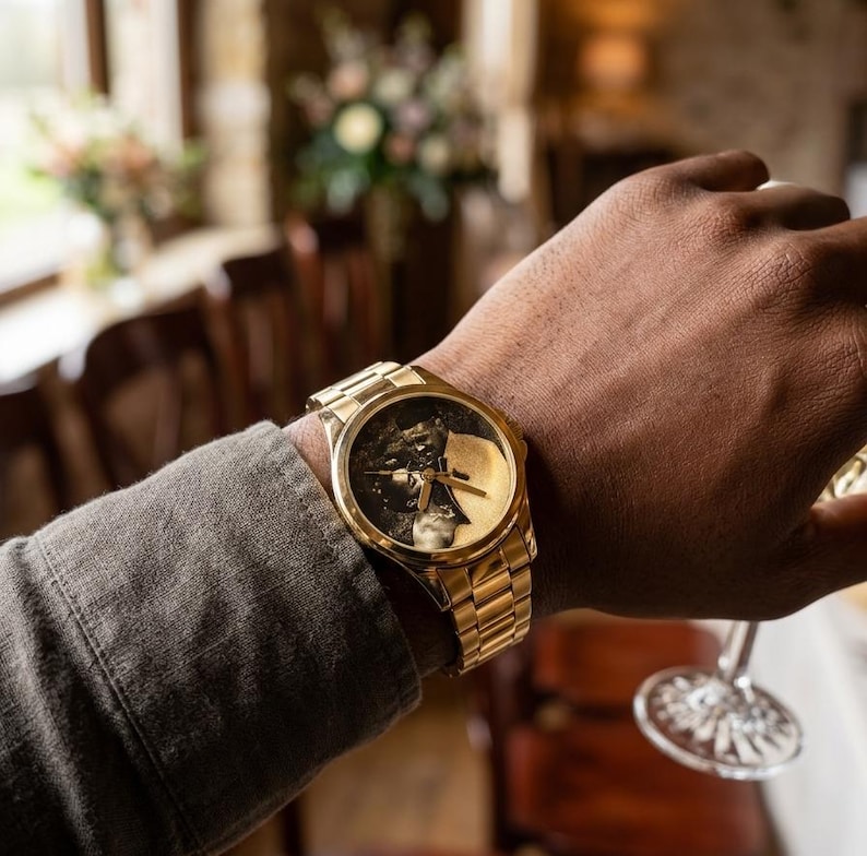 Puede incluir: Un reloj de pulsera dorado con una esfera detallada que presenta una imagen en blanco y negro. El reloj tiene una correa dorada y se lleva en la mu&ntilde;eca de una persona. El reloj es el punto focal, con un fondo borroso.