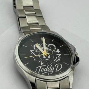 May include: A silver wristwatch with a black face featuring a bear graphic wearing sunglasses and the text "Teddy D". The watch has a silver band and gold-colored hands.