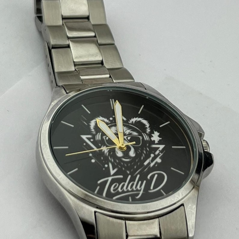 May include: A silver wristwatch with a black face featuring a bear graphic wearing sunglasses and the text "Teddy D". The watch has a silver band and gold-colored hands.